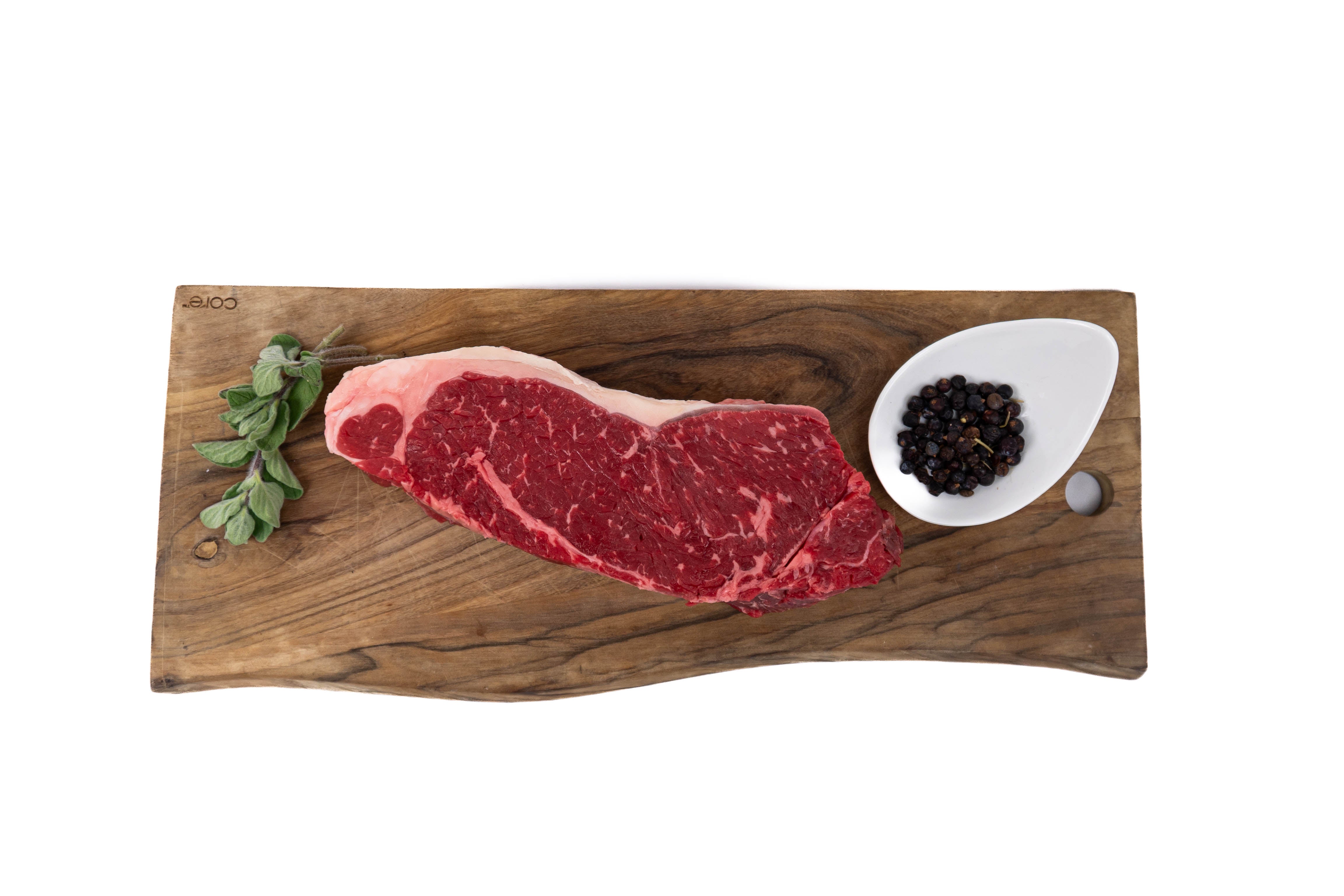 Raw steak on a wooden cutting board with a small bowl of peppercorns and a sprig of rosemary.