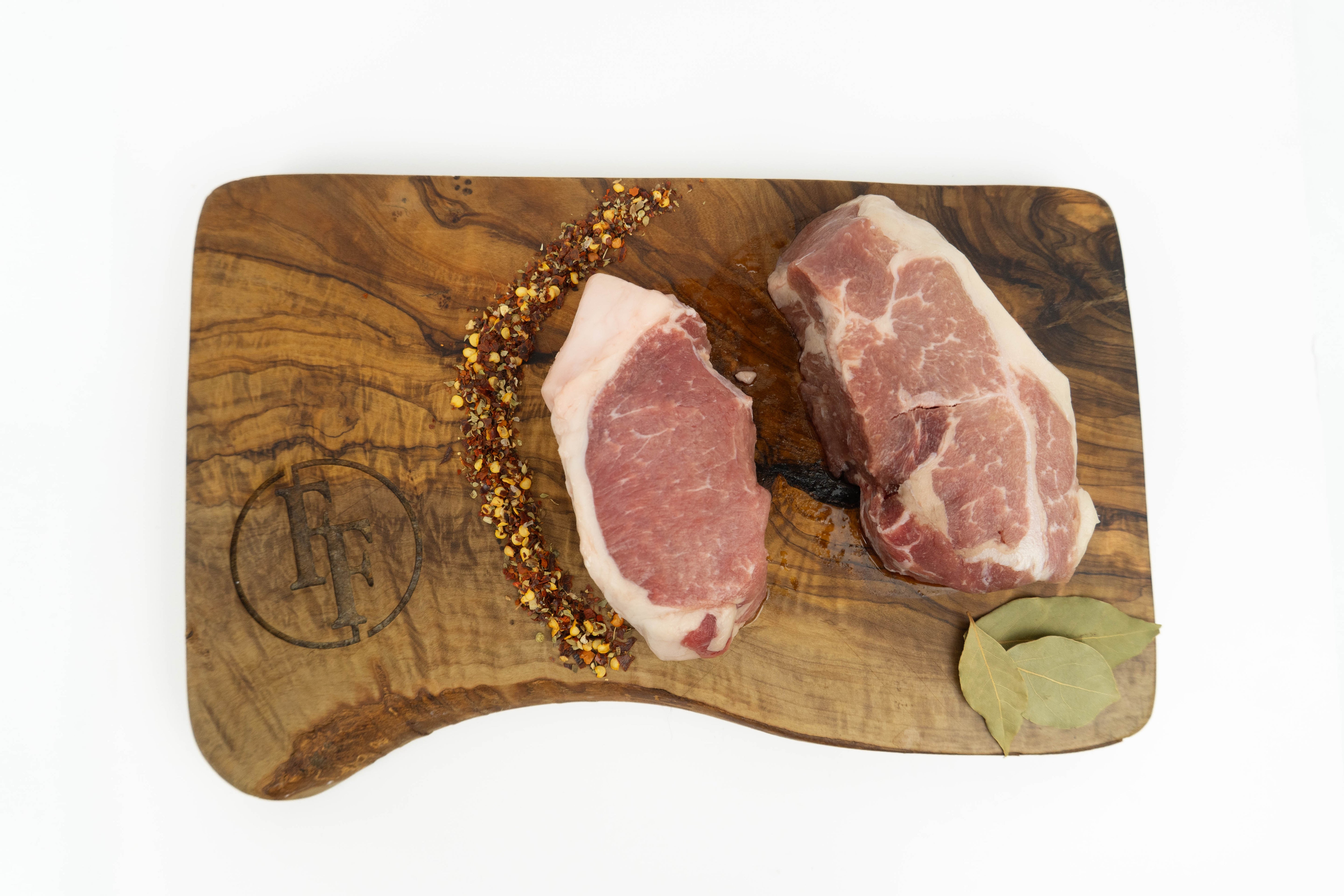 Two raw wild boar chops chops with visible marbling rest on a wooden cutting board, garnished with a line of dried peppercorns and a bay leaf, photographed from above on a white background.