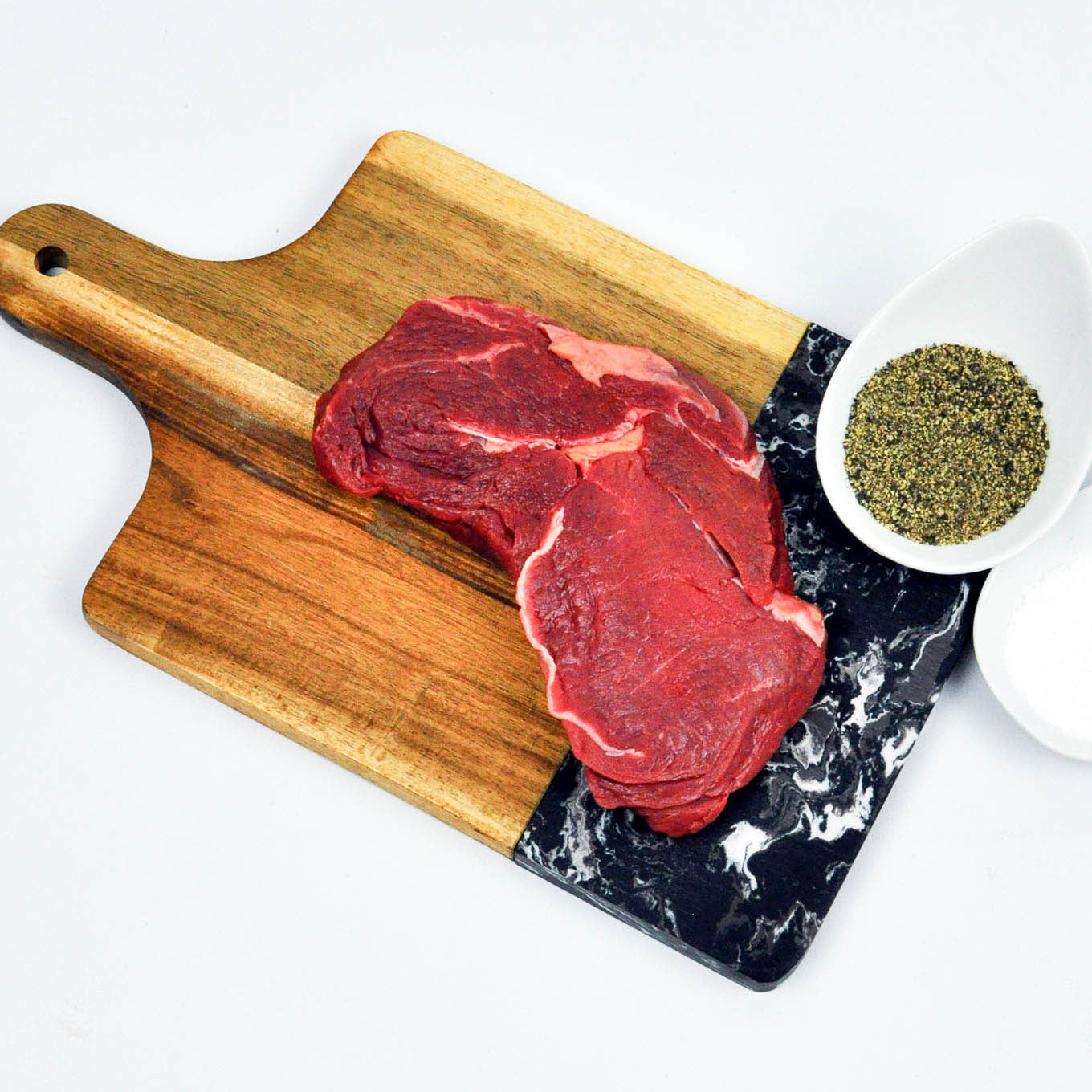 Raw steak on a wooden cutting board with a small bowl of herbs on a white background