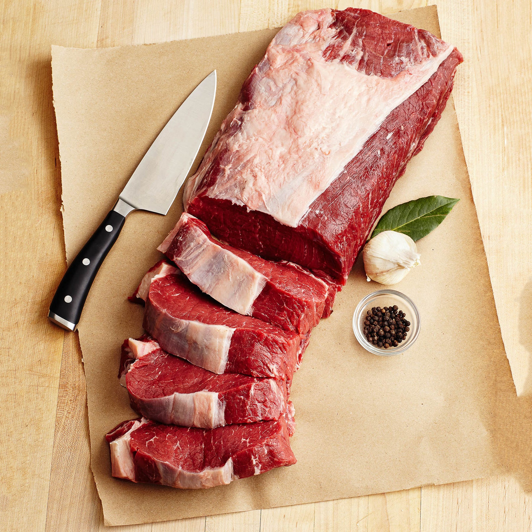 Raw beef with sliced pieces on brown paper, knife, garlic, and peppercorns.
