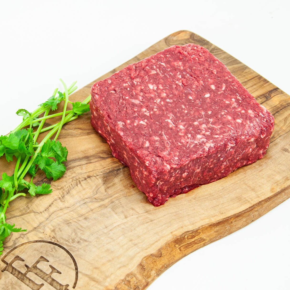 Block of ground meat on a wooden cutting board with a brand logo, surrounded by fresh greens.