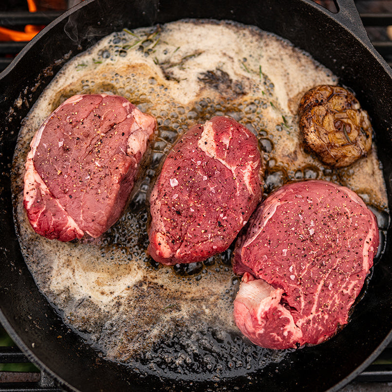 Three grassfed beef tenderloin steaks are cooking on a grill, shown with visible grill marks and smoke.