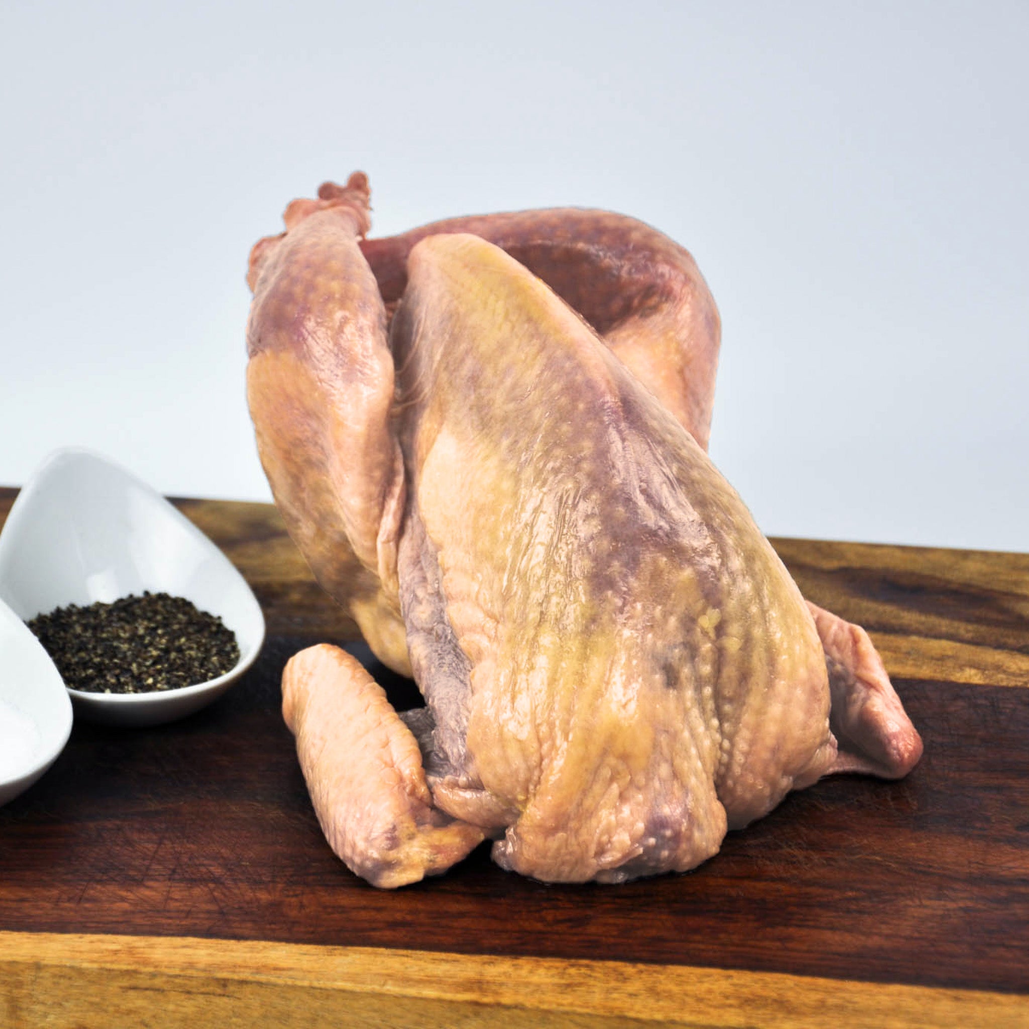 Raw guinea hen on a wooden cutting board with black pepper in a bowl.