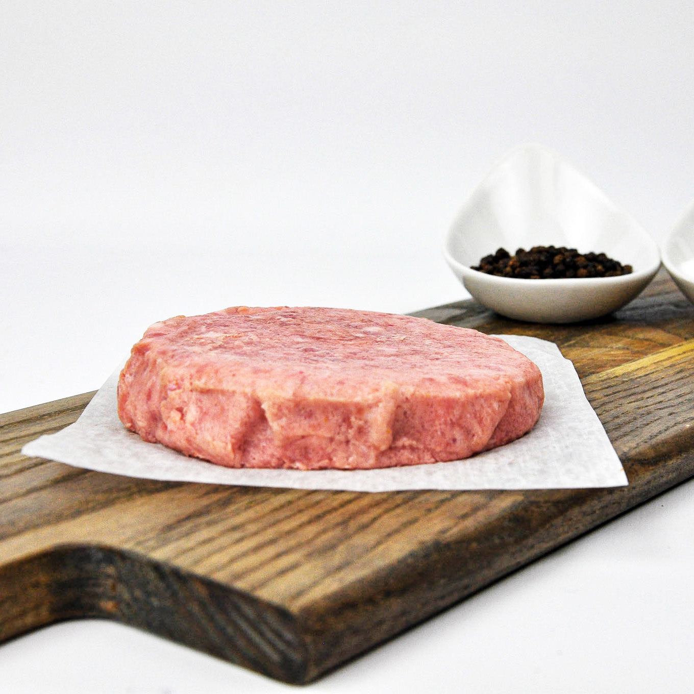 Raw hamburger patty on a wooden cutting board with a small bowl of spices in the background.