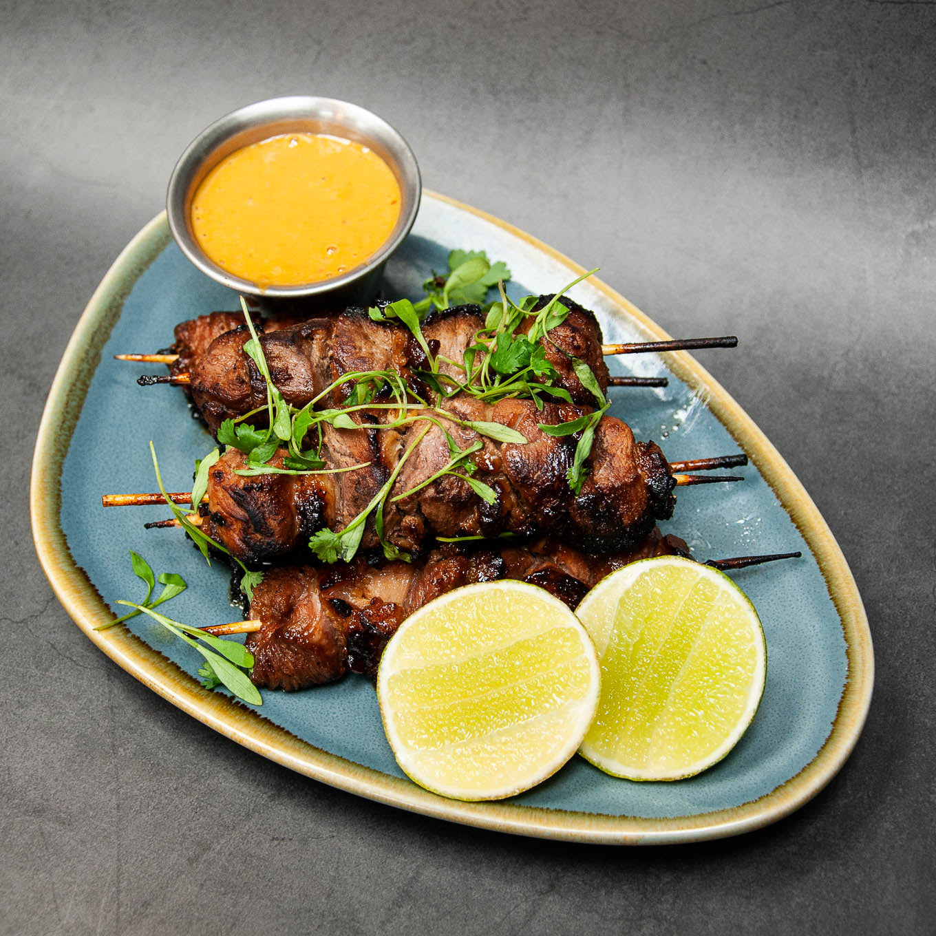 Skewers of grilled meat with lime wedges and a dipping sauce on a gray background