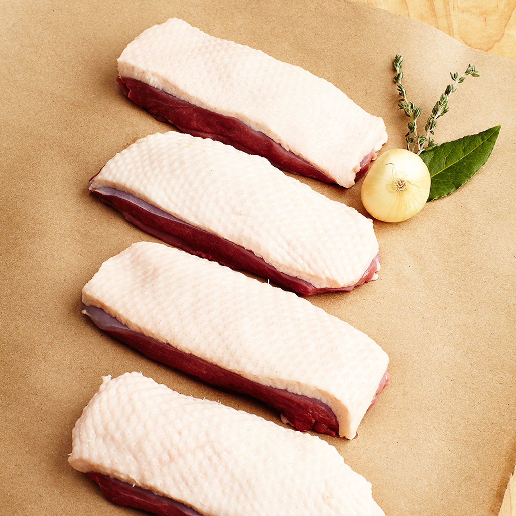 Four pieces of raw duck breast on a brown surface with a lemon and herbs.