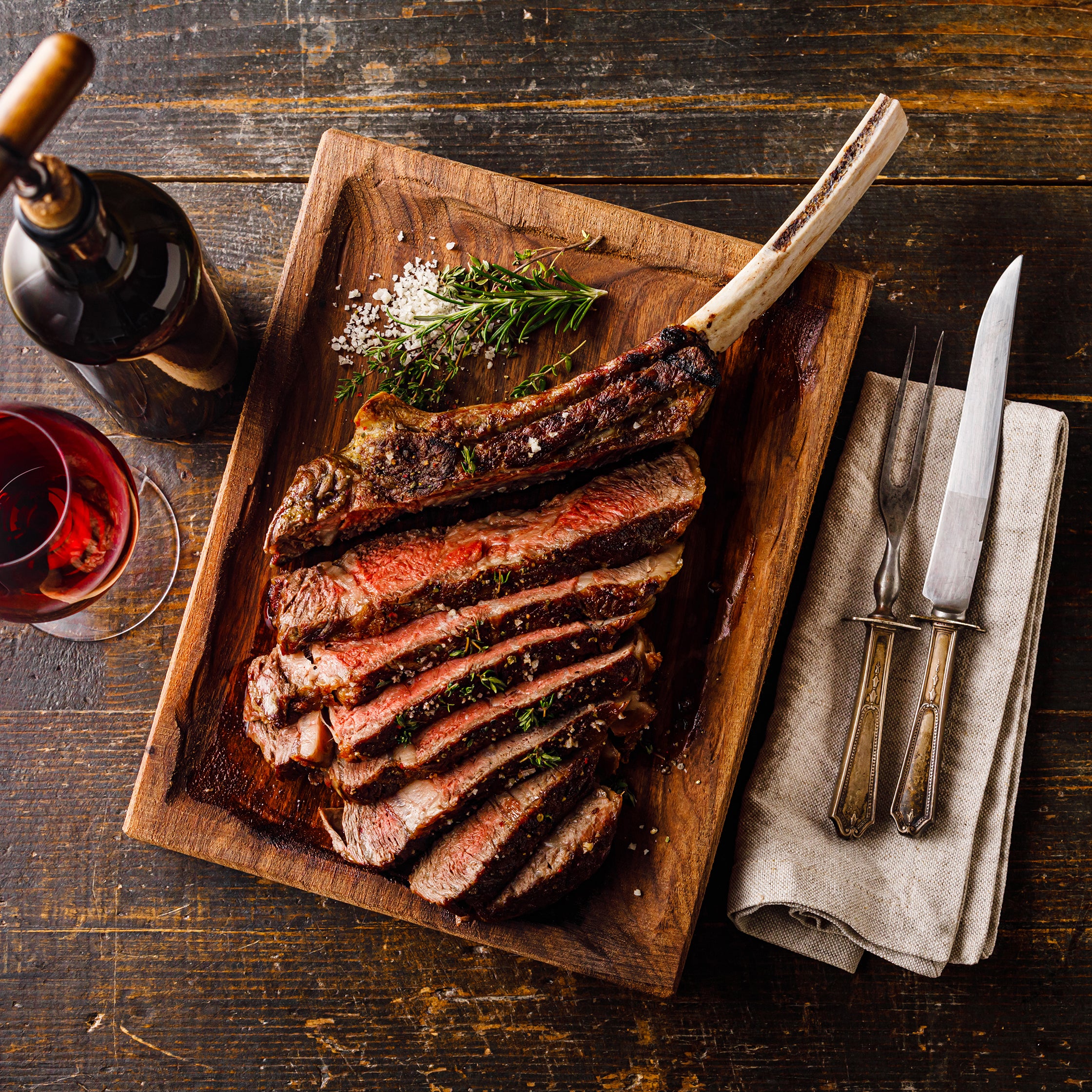 Sliced steak on a wooden platter with a bottle of wine and glasses on a rustic wooden table.