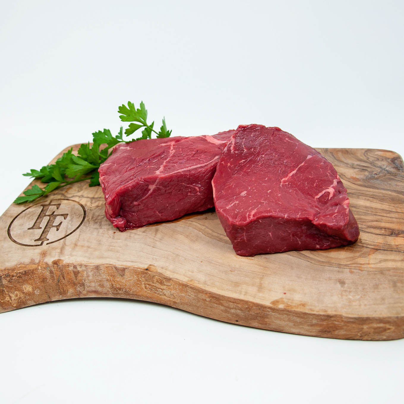 Two raw steaks on a wooden cutting board with a brand logo, surrounded by fresh parsley.
