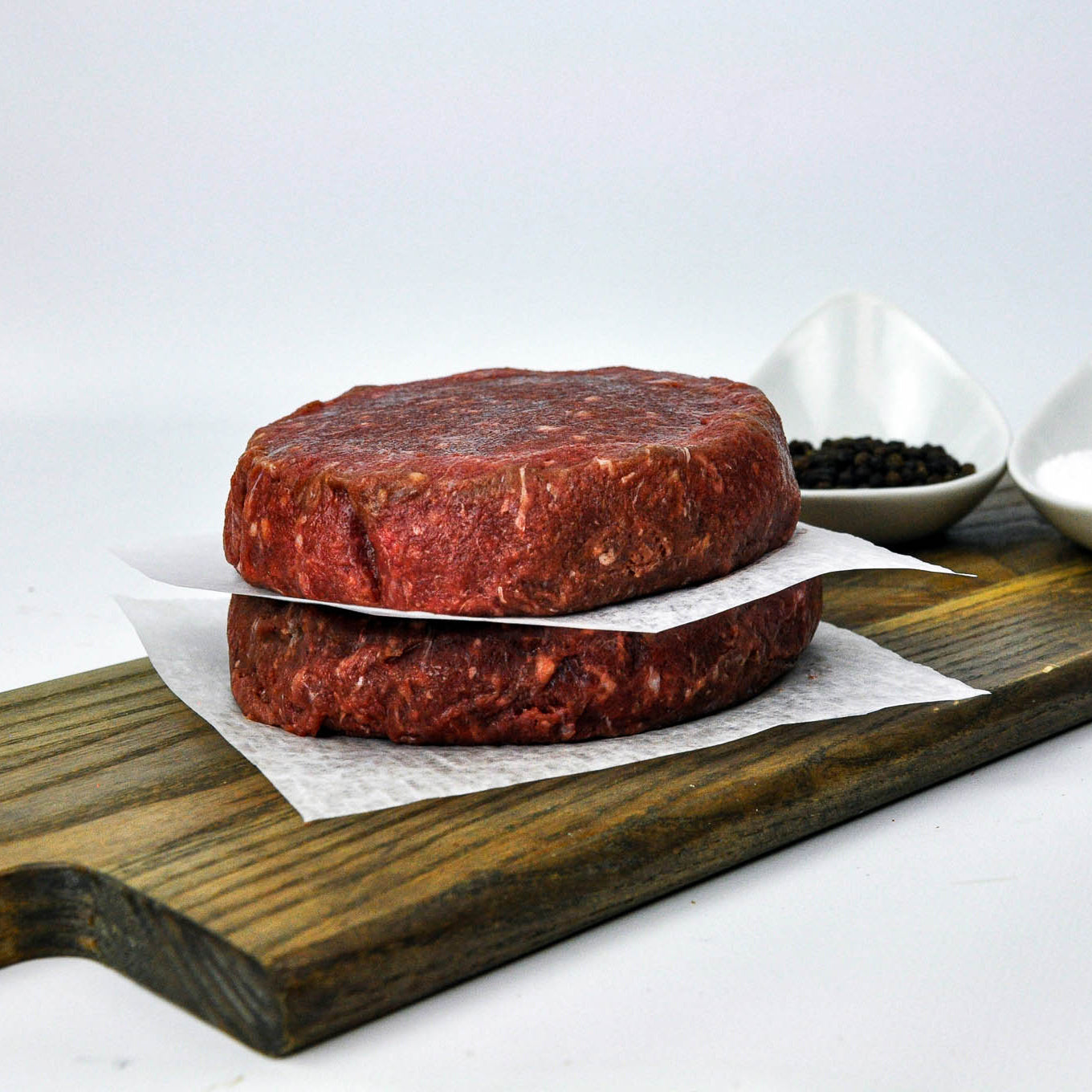 Two raw beef patties on a wooden cutting board with a white background