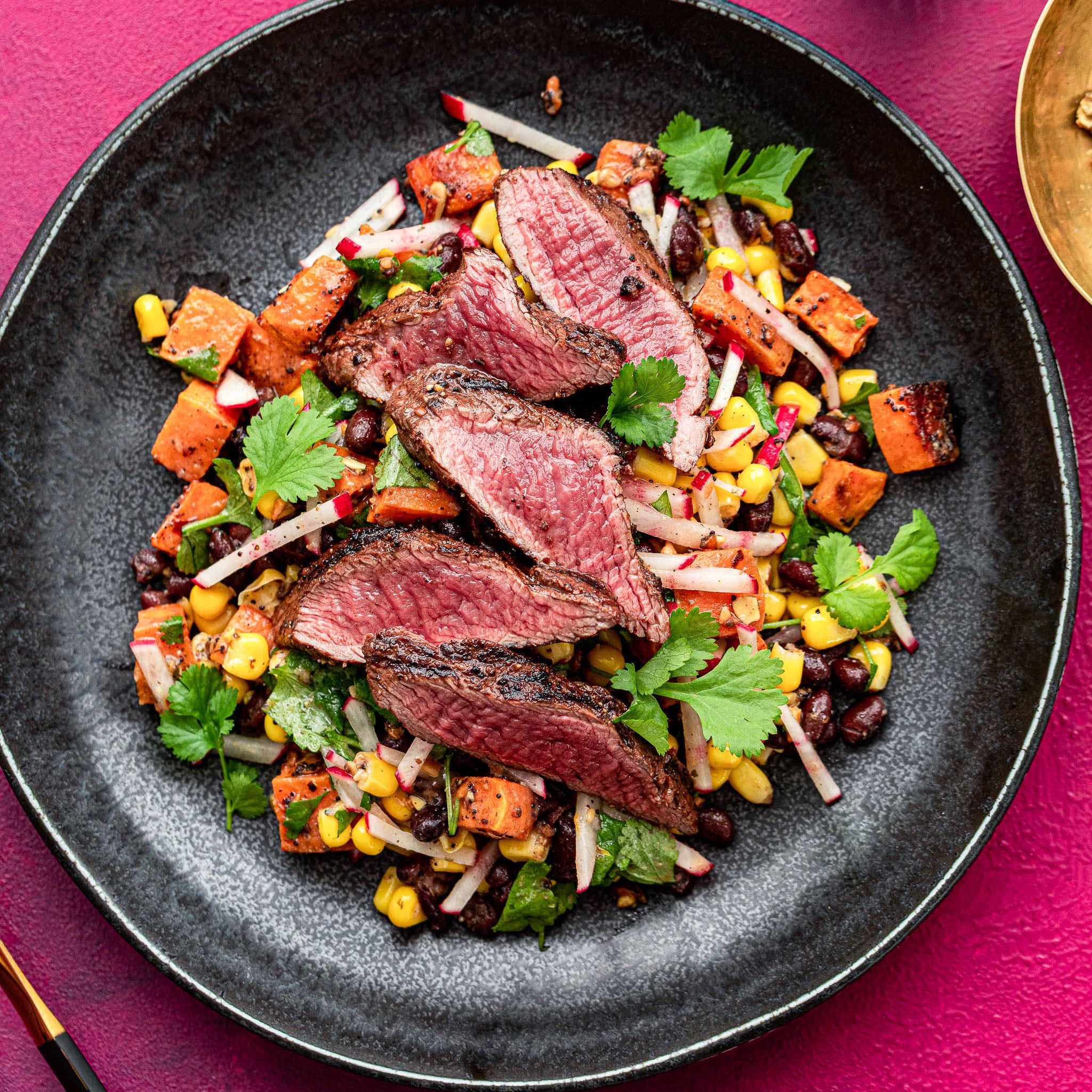 Plated dish of sliced steak with a colorful side salad on a pink background