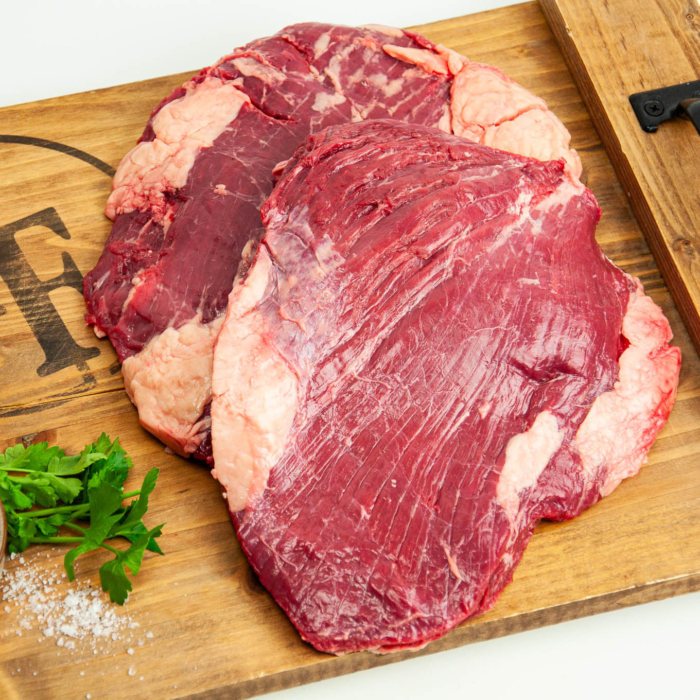 Two raw steaks on a wooden cutting board with herbs and salt.