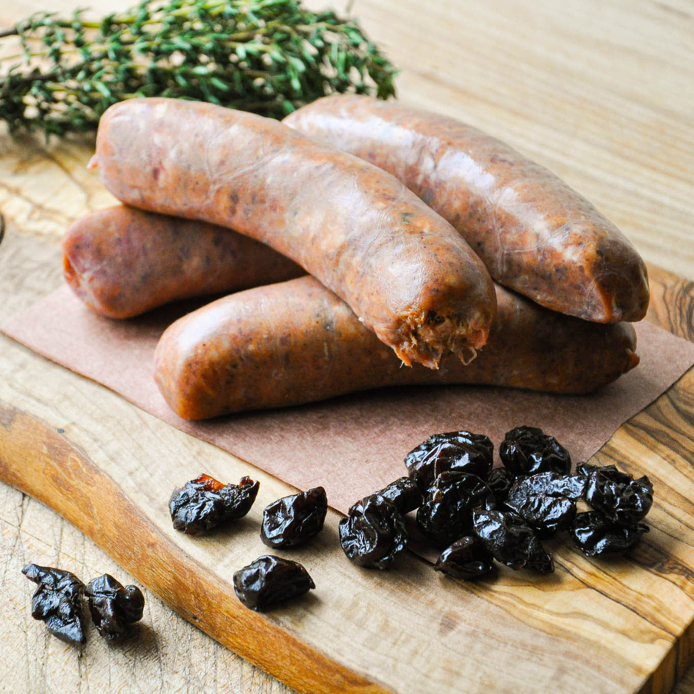 Three raw sausages on a wooden cutting board with dried plums and herbs.