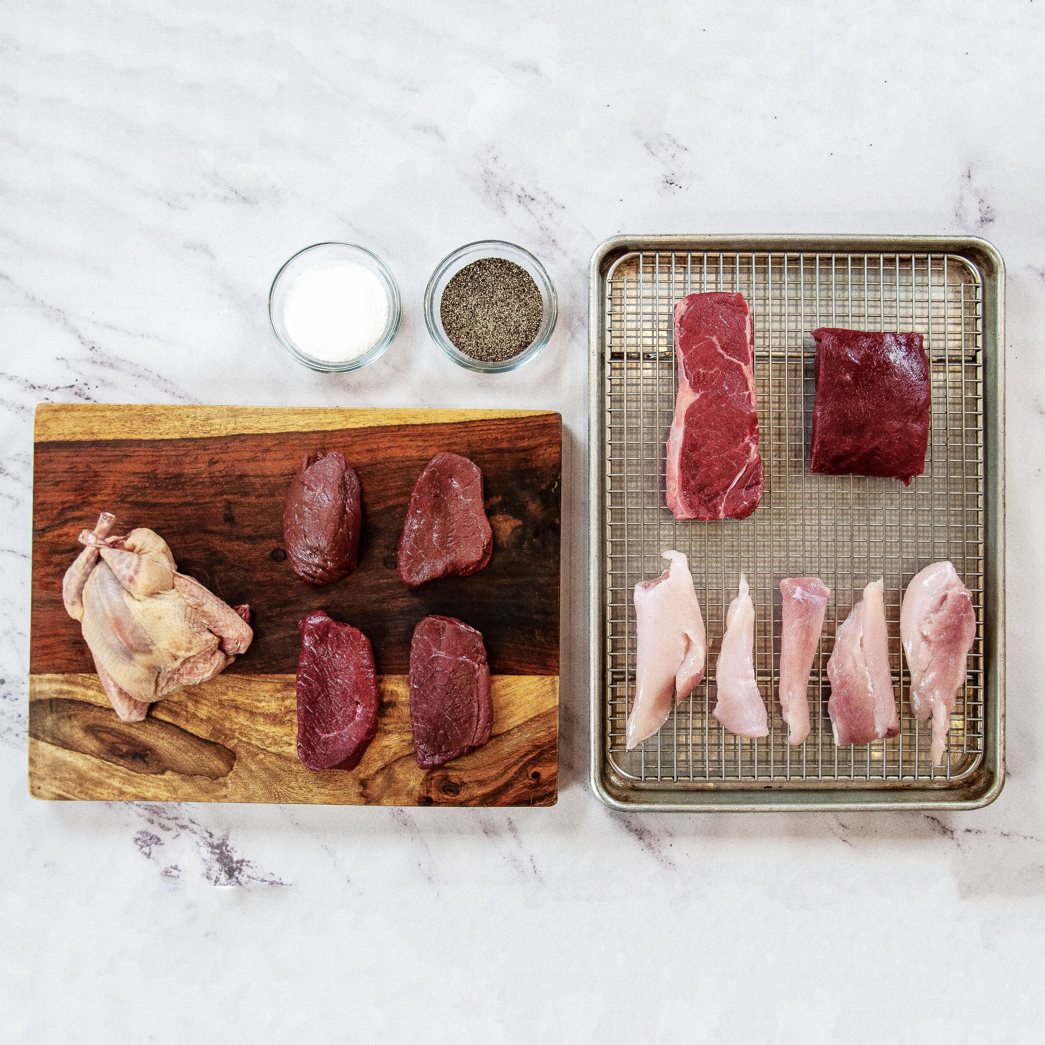 Raw chicken and beef on a wooden cutting board and metal tray with salt and pepper shakers.