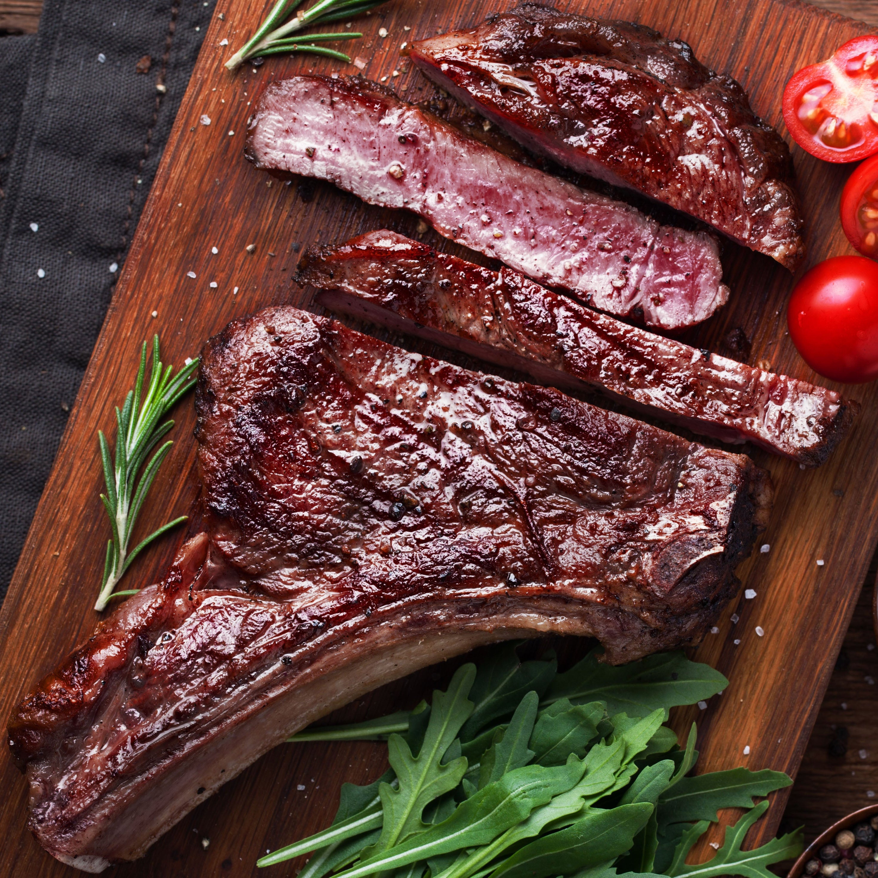 Grilled steak on a wooden cutting board with tomatoes and herbs on a rustic wooden surface.