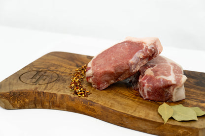 Two raw wild boar strip steaks with visible fat marbling, arranged on a wooden cutting board with red pepper flakes, bay leaves, and an engraved logo, photographed on a clean white background.