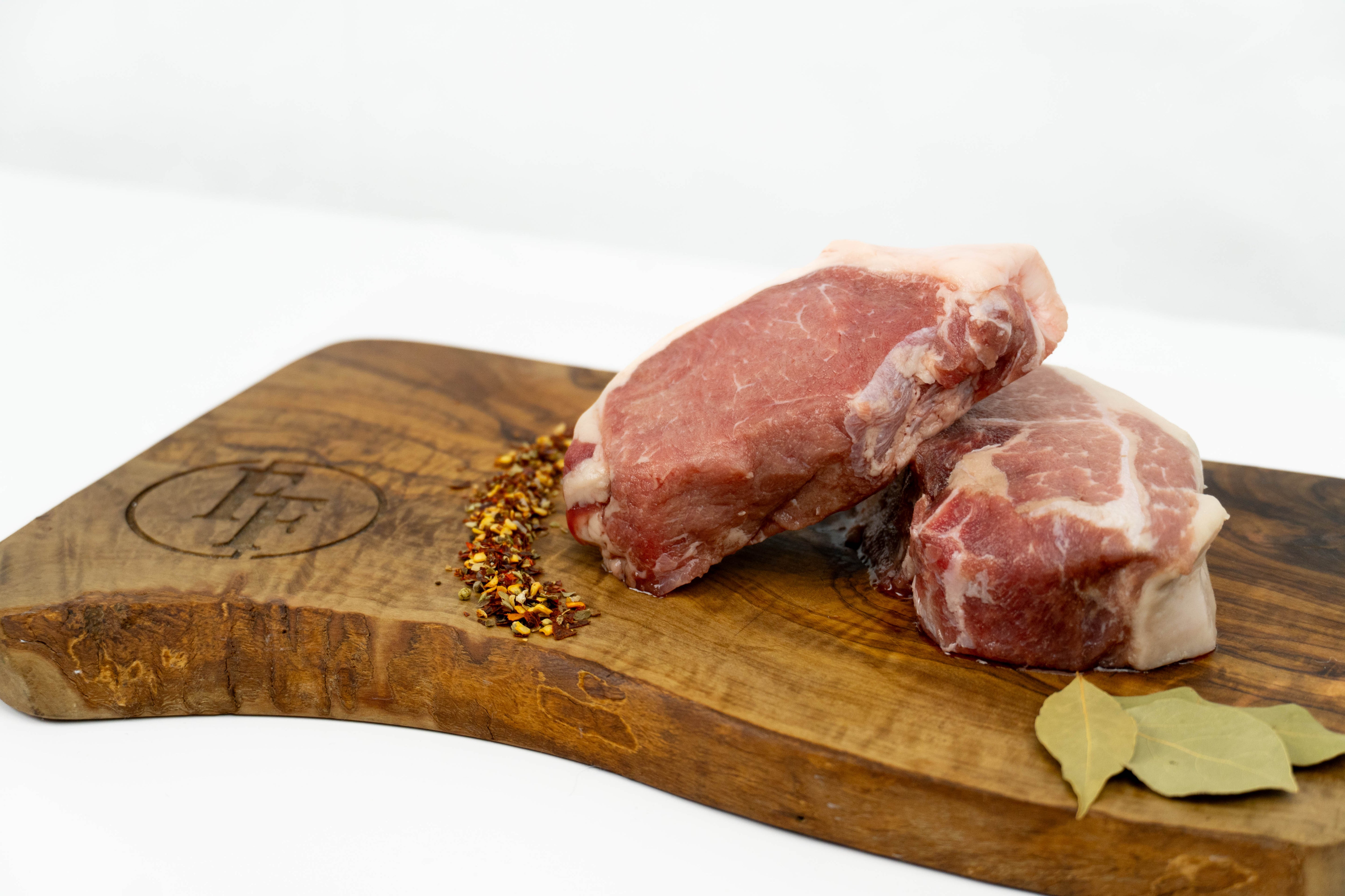 Two raw wild boar strip steaks with visible fat marbling, arranged on a wooden cutting board with red pepper flakes, bay leaves, and an engraved logo, photographed on a clean white background.