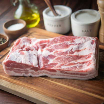Raw pork belly on a wooden cutting board with salt and pepper containers in the background.