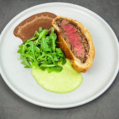 Plated dish with a sliced meat Wellington, green sauce, and arugula on a white plate.