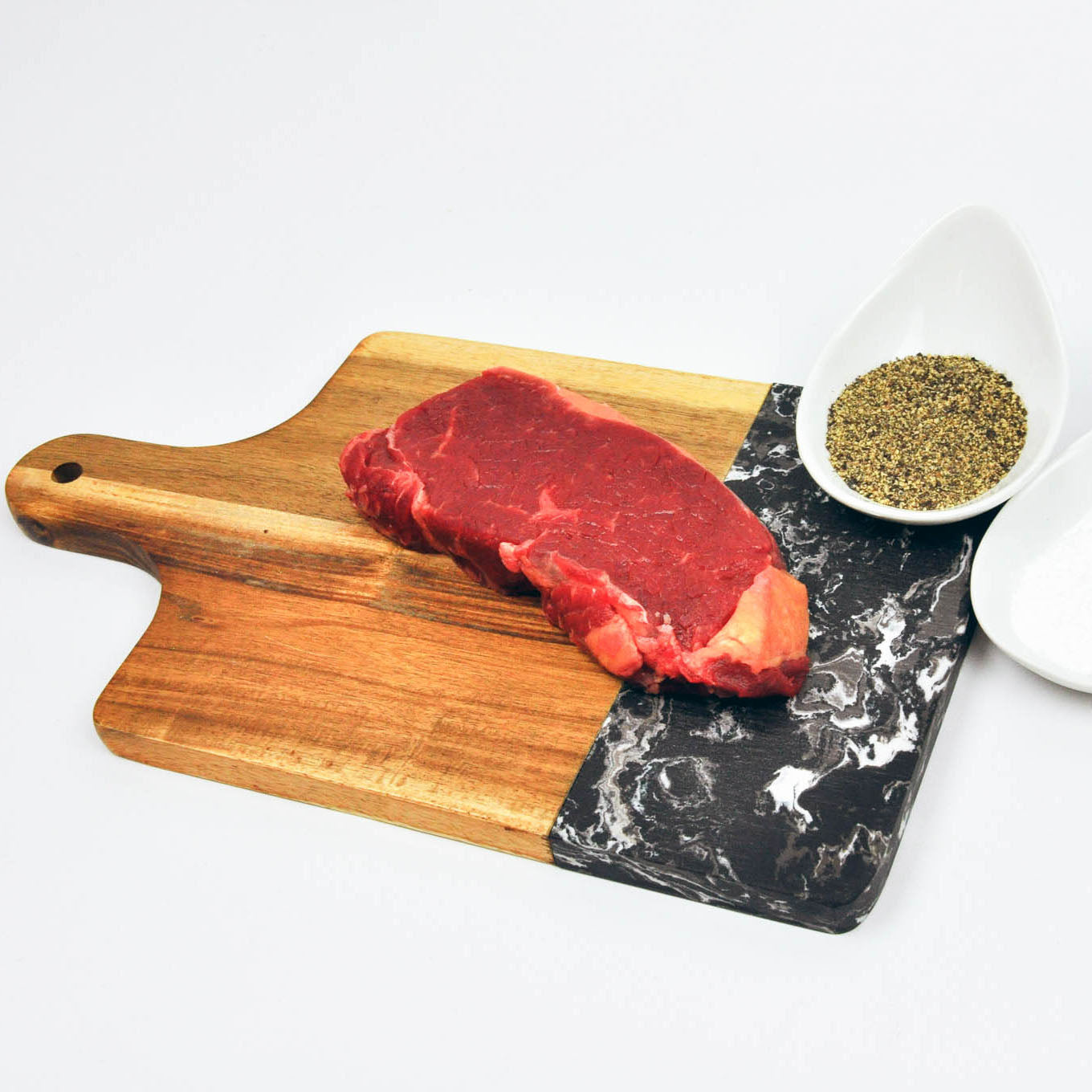 Raw steak on a wooden and marble cutting board with a bowl of seasoning.