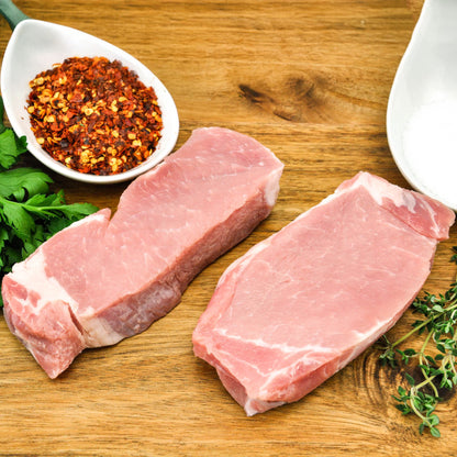 Two raw pork chops on a wooden cutting board with red pepper flakes and herbs.
