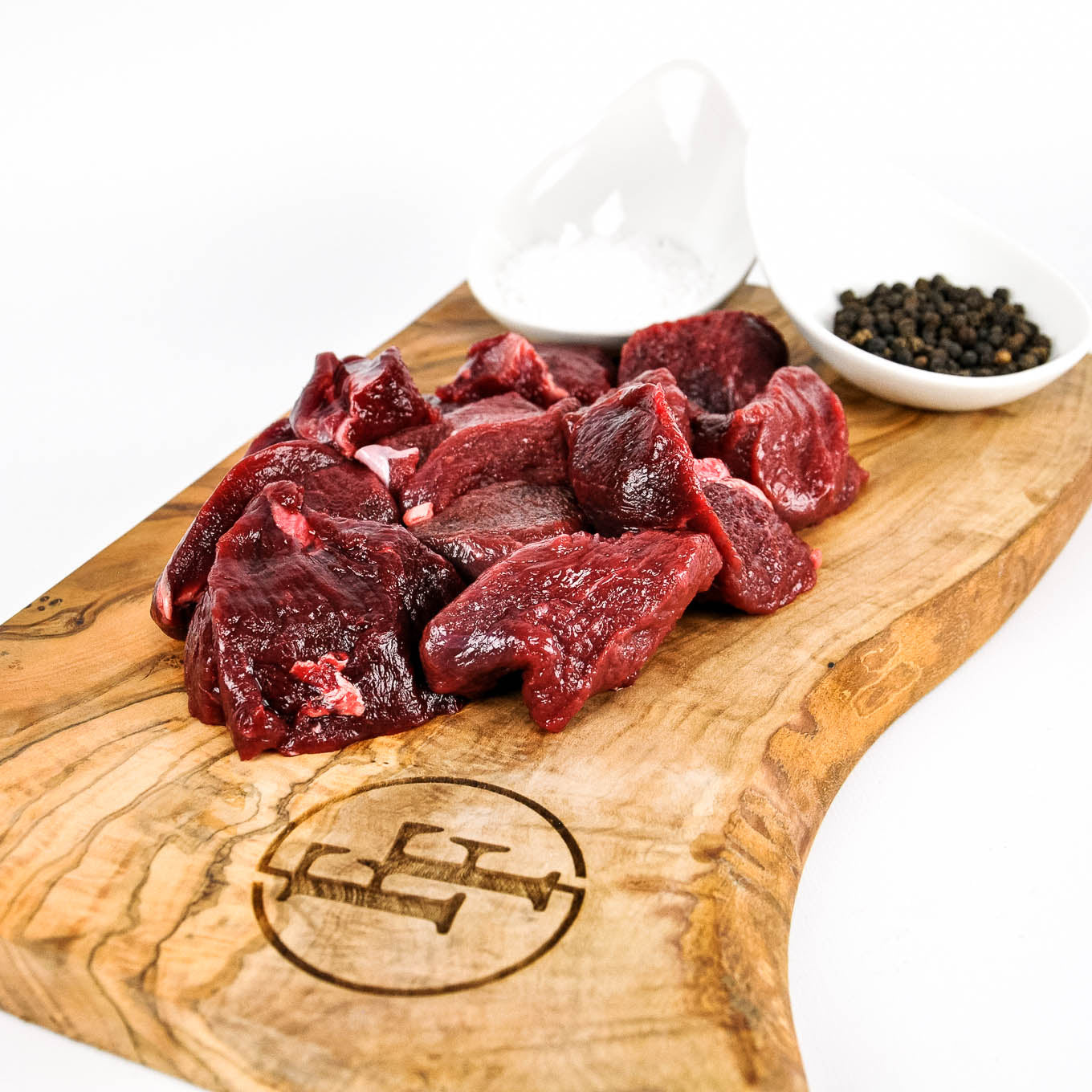 Raw beef chunks on a wooden cutting board with a logo, surrounded by small bowls of spices.