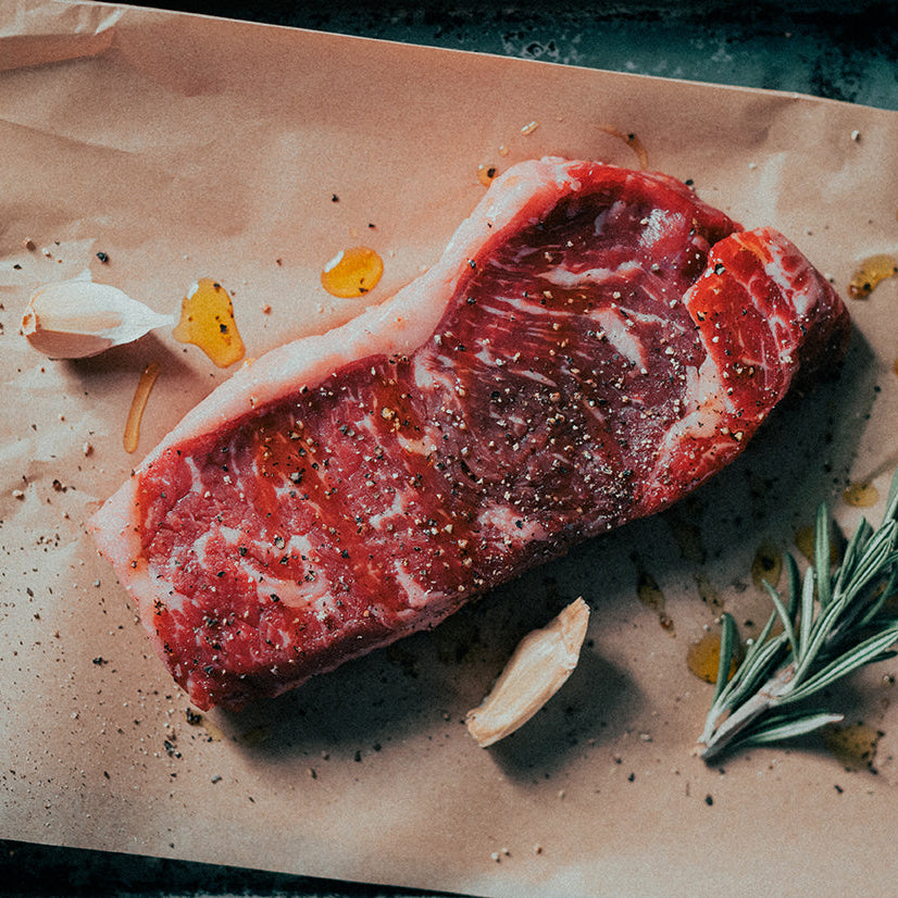 A 12oz grassfed beef striploin steak on a piece of brown butcher paper with a sprig of rosemary and a clove of garlic next to it.