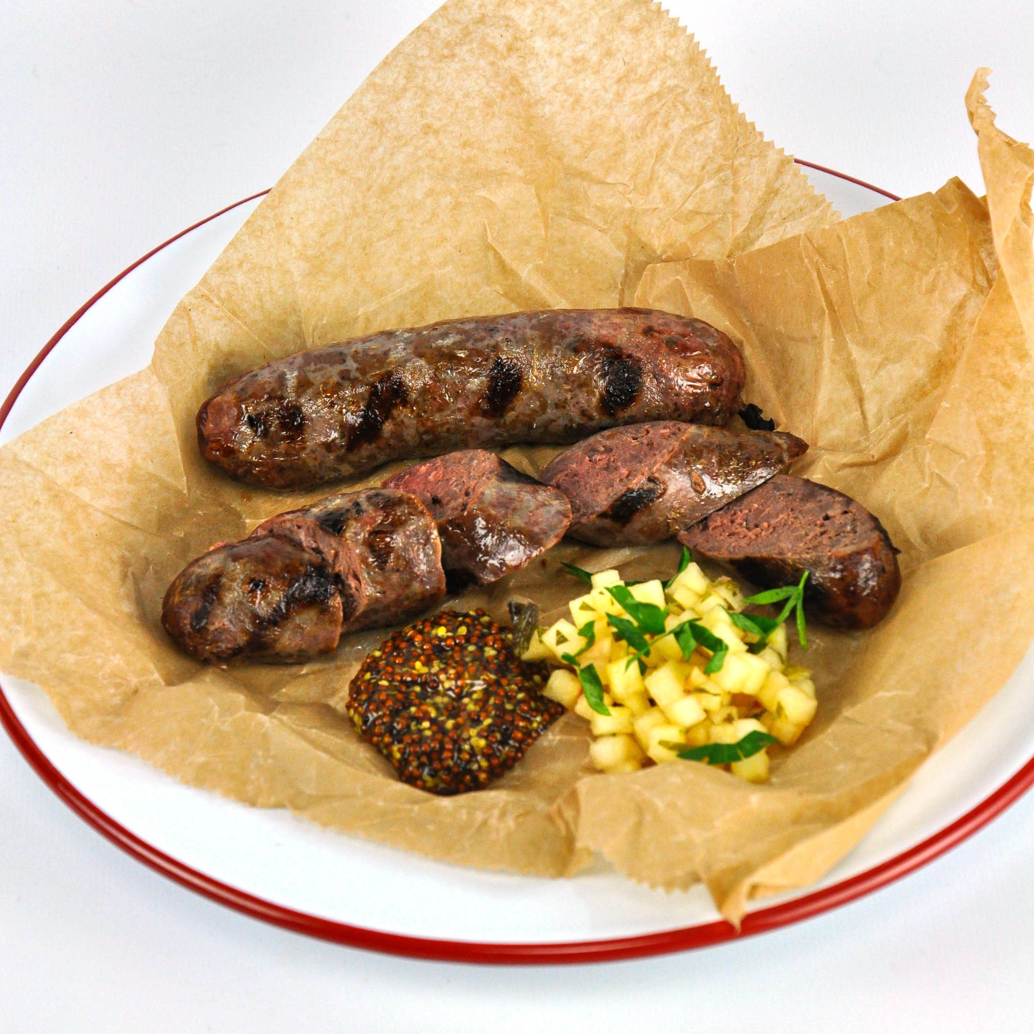 Grilled sausages on brown paper with a side of yellow food and red-rimmed plate