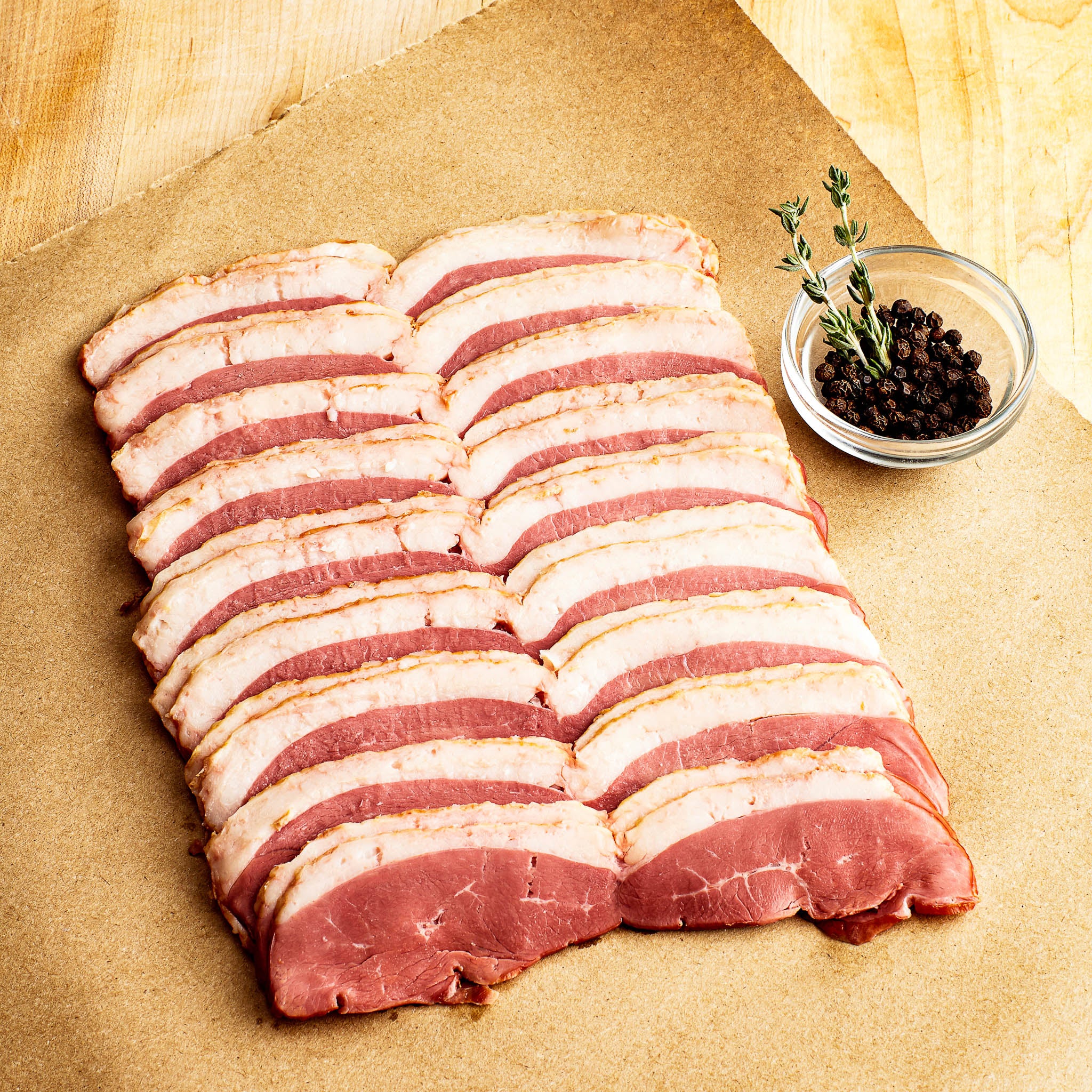 Raw bacon slices on a wooden surface with a small bowl of black peppercorns.