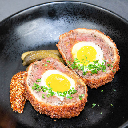 Two scotch eggs on a black plate with a side of pickles.