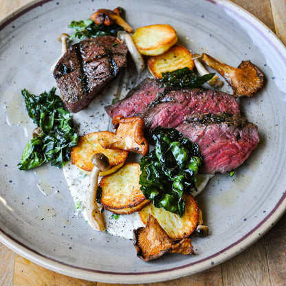 Plated dish of sliced steak with greens and mushrooms on a rustic plate.