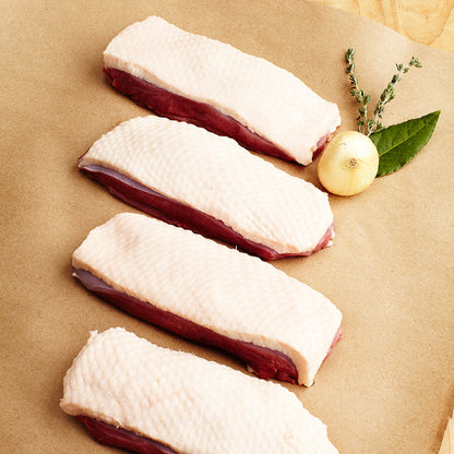 Four pieces of raw duck breast on a brown surface with a lemon and herbs.