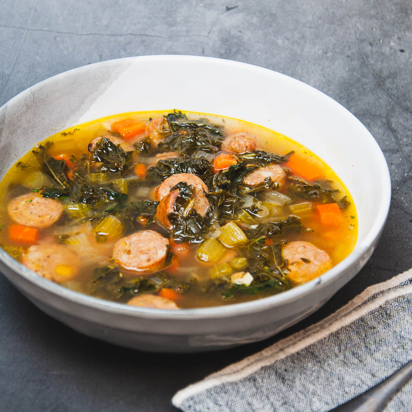 Bowl of soup with sausages, greens, and carrots on a gray surface