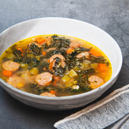Bowl of soup with sausages, greens, and carrots on a gray surface