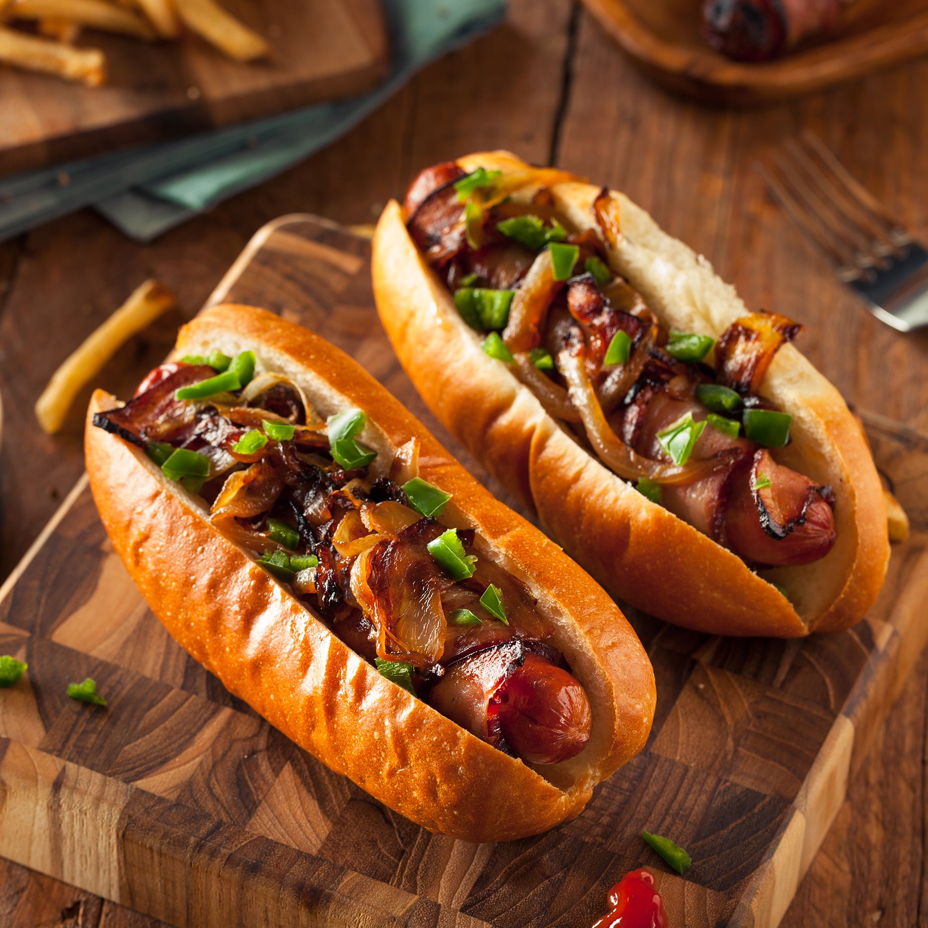 Two hot dogs with onions and green onions on a wooden cutting board.