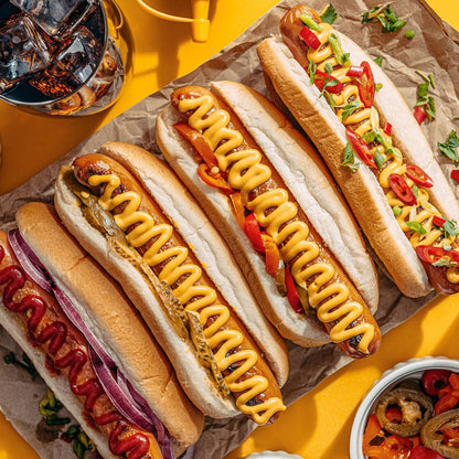 Three hot dogs with mustard, onions, and peppers on a yellow surface with a drink in the background.