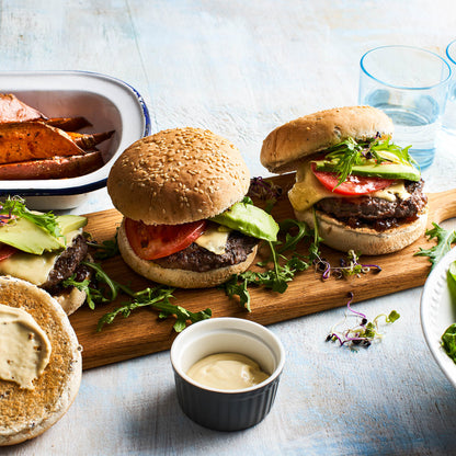 Three burgers on a wooden board with a side of sweet potato wedges and a glass of water.