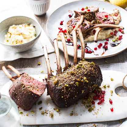 Two roasted lamb racks on a marble cutting board with side dishes in the background.