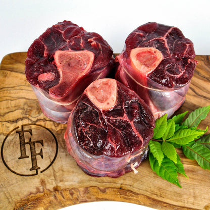 Three raw beef bones on a wooden cutting board with a brand logo, surrounded by green leaves.