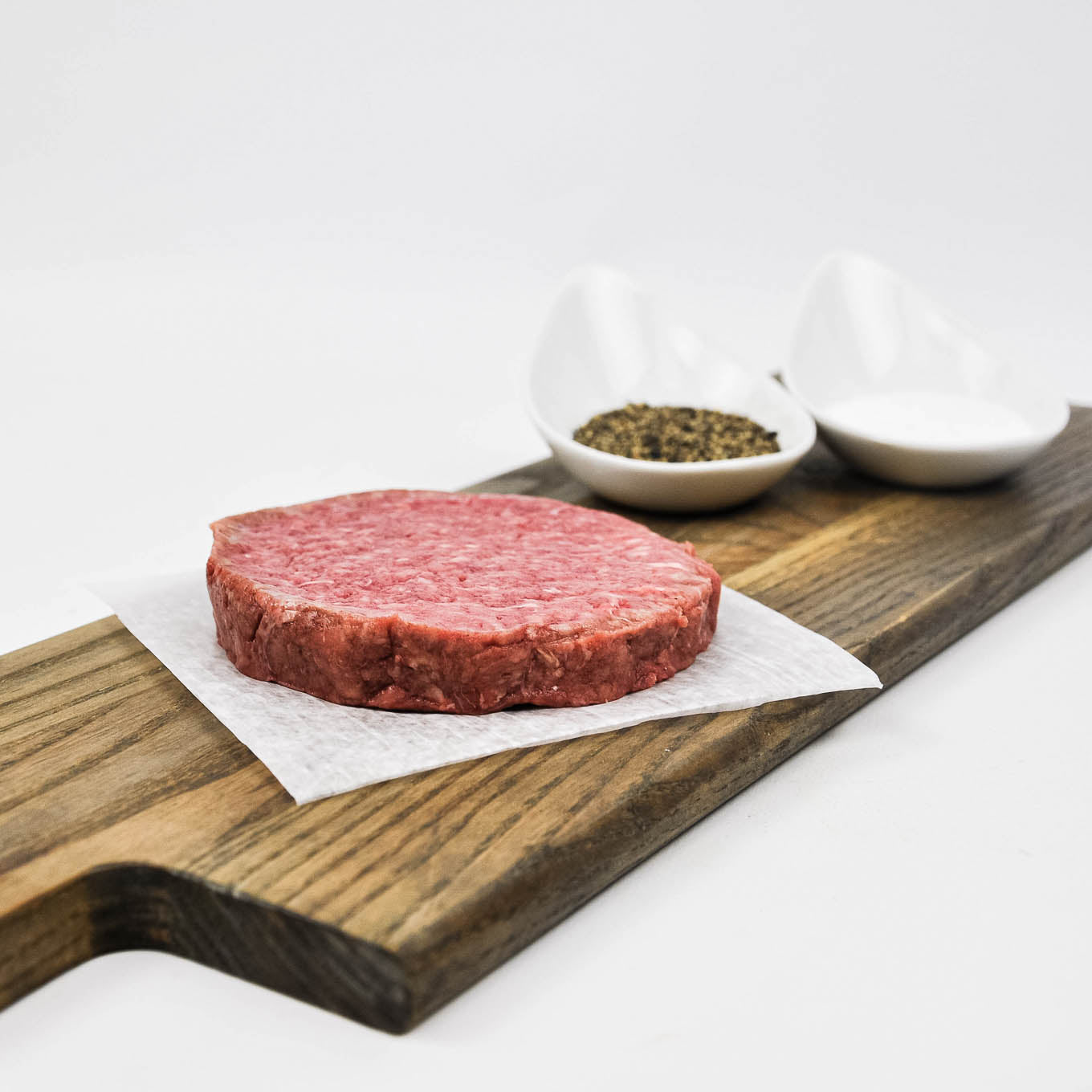 Raw hamburger patty on a wooden cutting board with small bowls of spices in the background.
