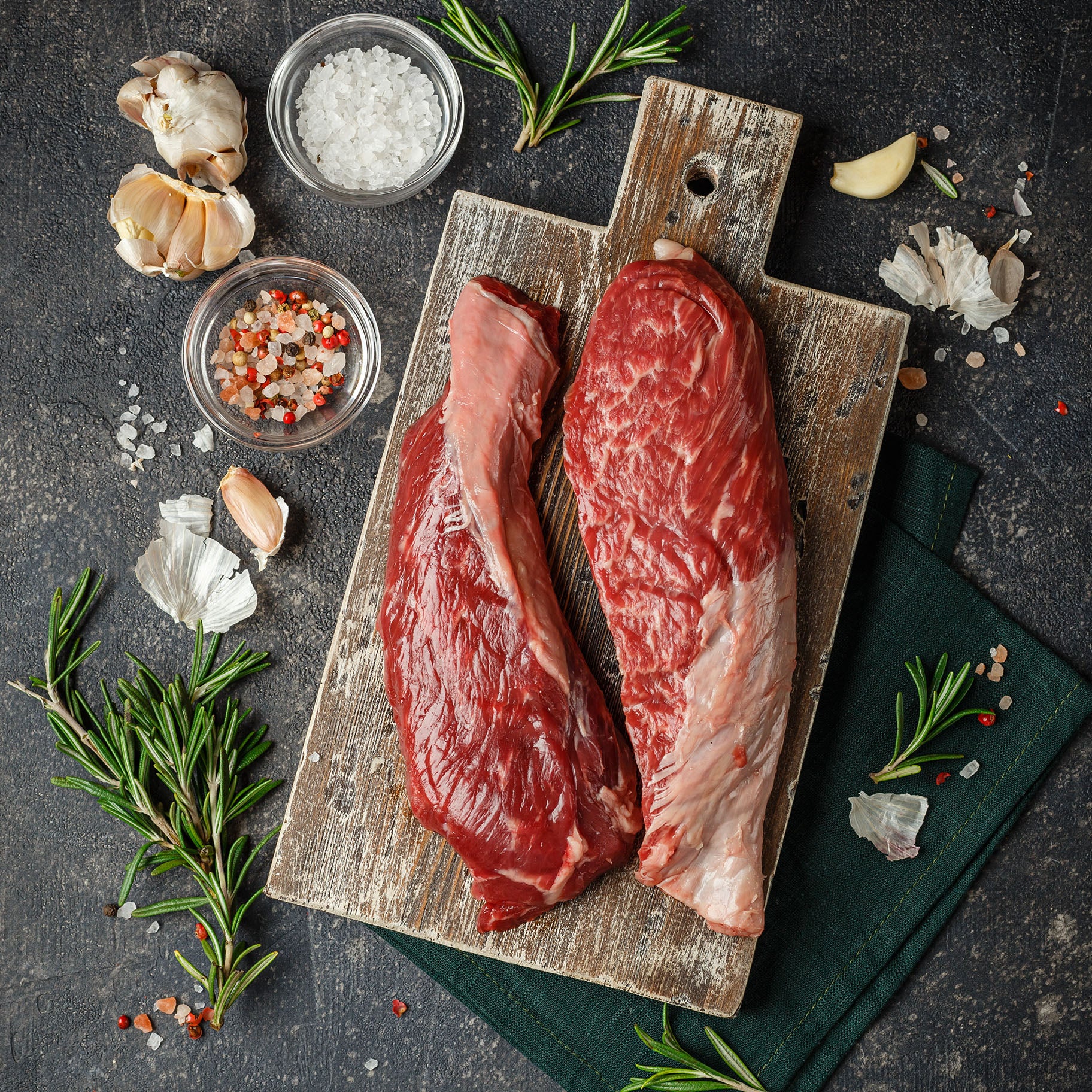 Two raw steaks on a wooden cutting board with herbs and spices on a dark surface