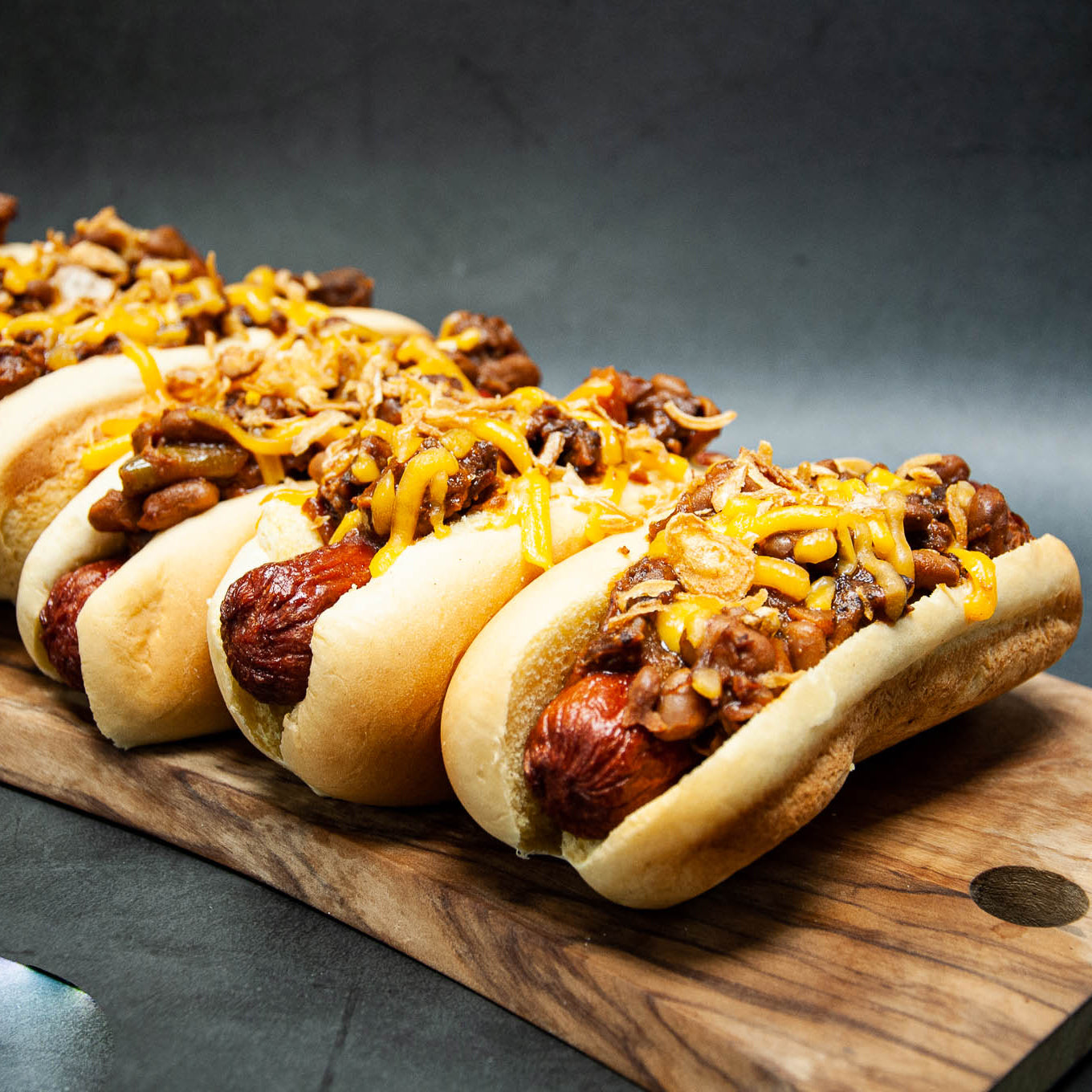 Three hot dogs with chili and cheese on a wooden board against a dark background