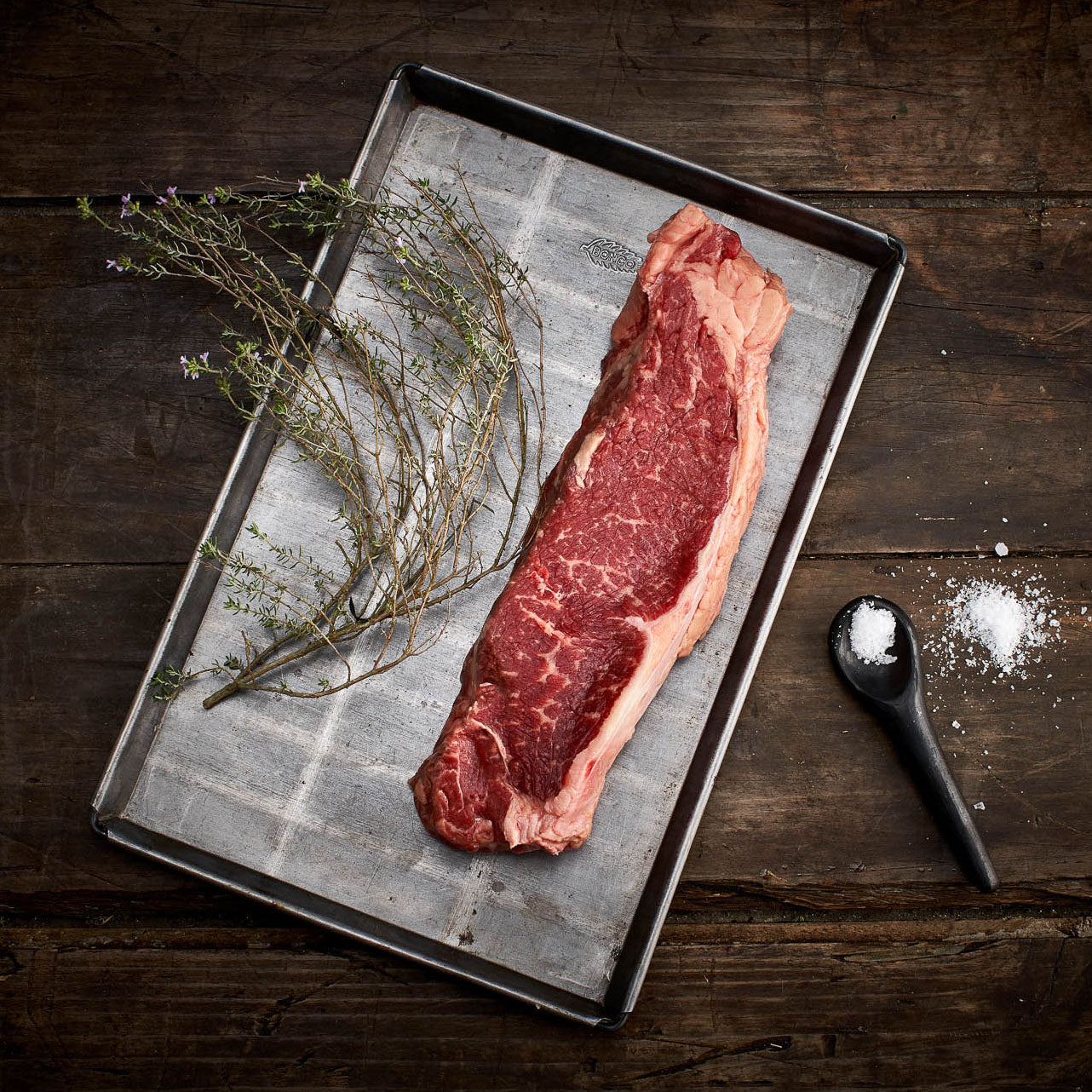 Raw steak on a metal tray with herbs and salt on a wooden surface