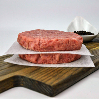 Two raw hamburger patties on a wooden cutting board with a small bowl of spices.