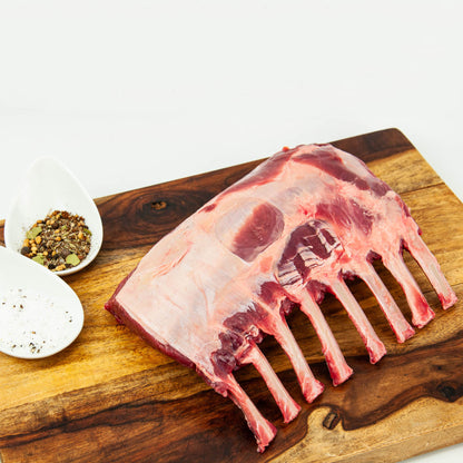 Raw lamb rack on a wooden cutting board with seasonings in the background