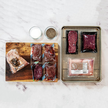 Assorted raw meat packages on a marble surface with a wooden cutting board and small containers of salt and pepper.